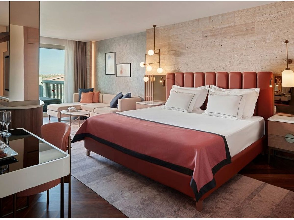 A suite bedroom with a terracotta upholstered headboard and a red bed runner, a mirrored dressing table and the suite's living room and balcony beyond.