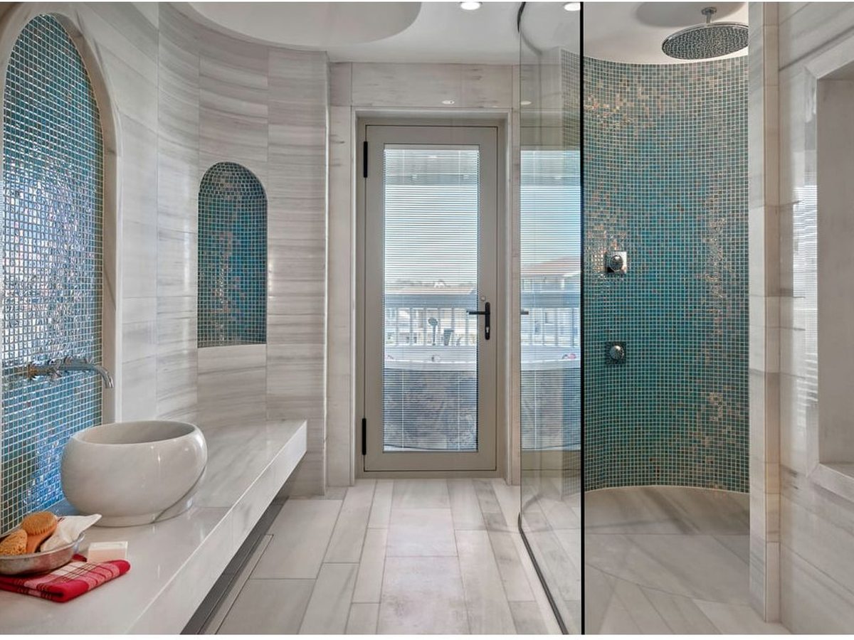 A bathroom lined in pale marble and blue-green mosaic with arched alcoves, a domed ceiling, a round stone basin and a glass door to a balcony.