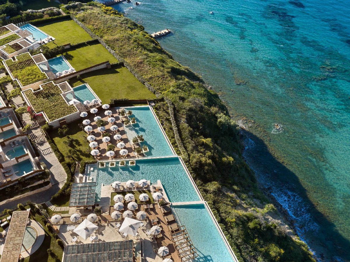Aerial view of three turquoise infinity pools on stepped terraces above a rocky shoreline, with rows of loungers and cream parasols between them.