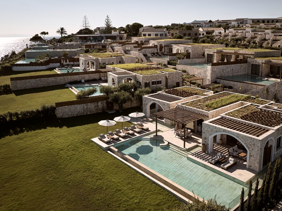 Aerial view of low stone buildings with planted turf roofs, private pools and mown lawns stepping down a hillside towards the sea.