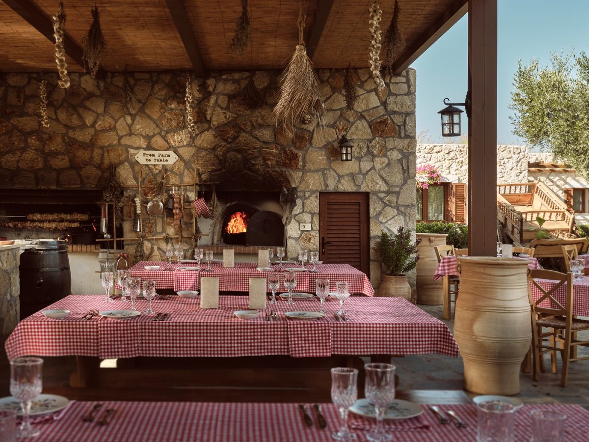 Covered stone dining room with a lit wood-fired oven, long tables under red gingham cloths, hanging dried herbs and large terracotta jars.
