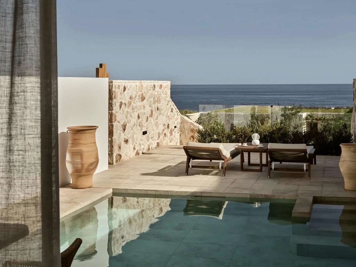 Small rectangular private pool on a stone terrace enclosed by rough stone walls, with a daybed, a terracotta amphora and the sea beyond.
