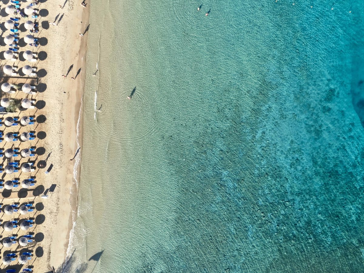 Aerial view of a long sandy beach lined with rows of blue parasols and sunbeds, with clear turquoise shallows running out to deeper water.