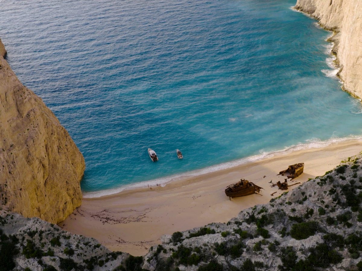 Aerial view of the rusting shipwreck on the sand at Navagio, at the foot of pale limestone cliffs, with two small boats anchored in turquoise water.