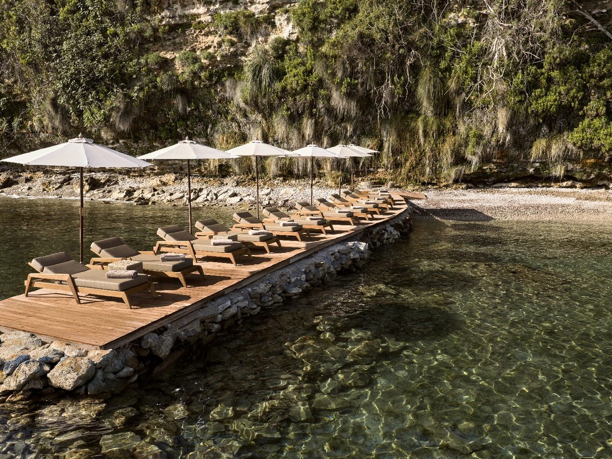 Wooden pier running out over clear turquoise water from a stone-edged shore, lined with timber sun loungers and white parasols.