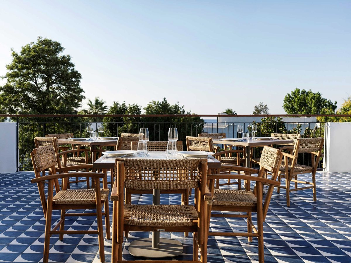 Square wooden tables and woven rattan armchairs set with glassware on a blue and white tiled terrace, behind a railing, with white rooftops and the sea beyond.