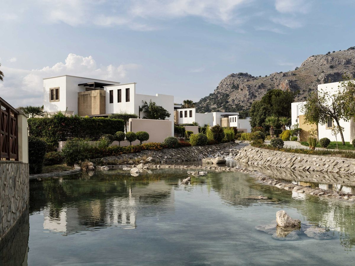 White flat-roofed accommodation blocks and clipped hedges above a wide stone-edged water channel, with a rocky hillside behind under a blue sky.