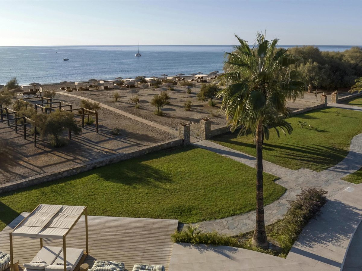 Mown lawns, stone-paved paths and a tall palm running down to a beach of straw parasols and timber pergolas, with a calm sea and a moored catamaran beyond.
