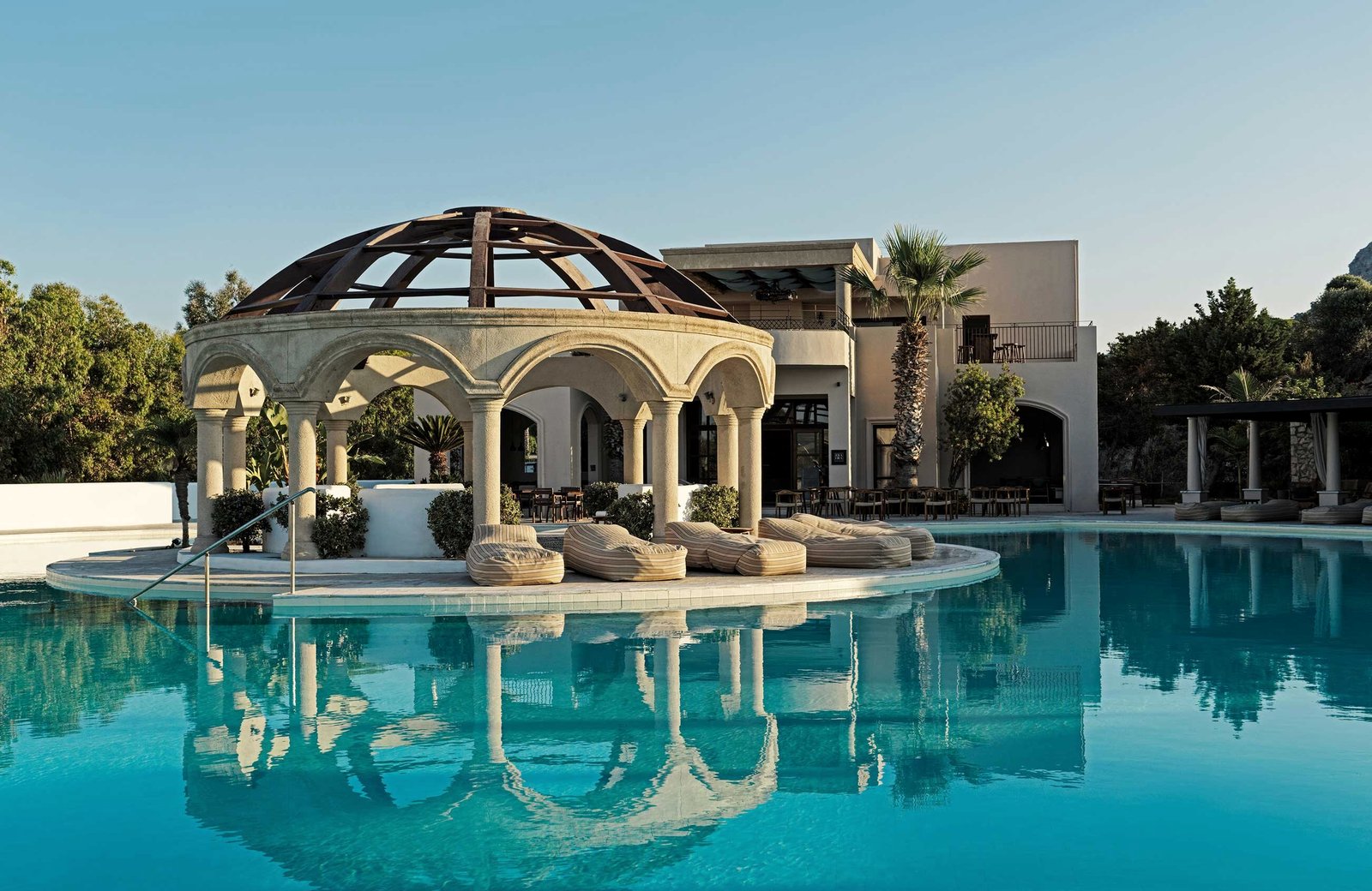 The resort's freeform turquoise pool, with a domed stone pavilion and cushioned loungers on an island at its edge and the white resort buildings behind.