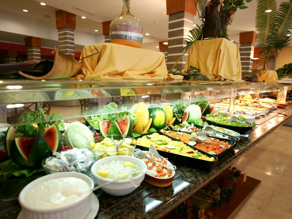 Cold buffet counter under a glass guard with composed salads, sliced vegetables, dressings and a carved watermelon.