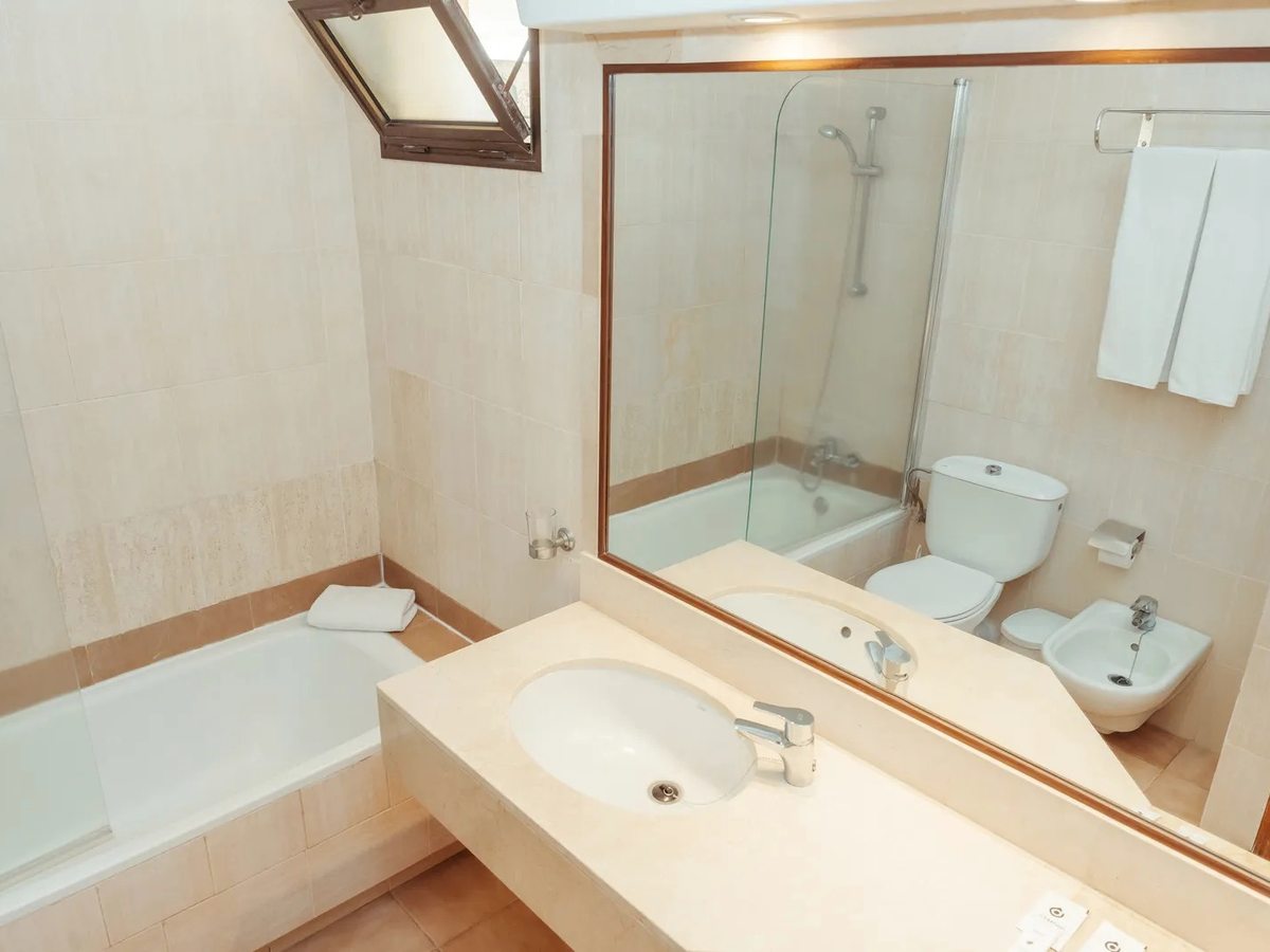 Bathroom with a full-size bath behind a glass screen, a wide vanity basin under a large mirror, a toilet and bidet, and cream tiling.