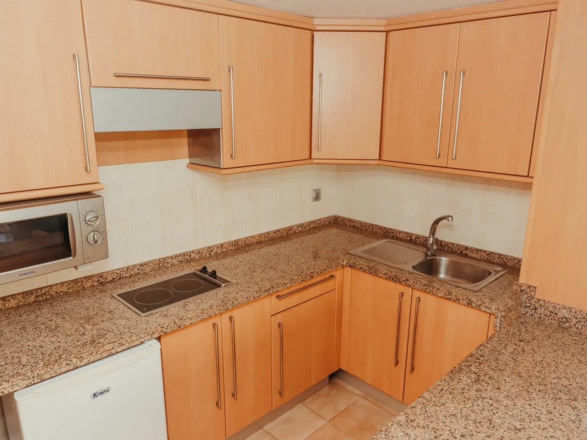 L-shaped kitchenette with pale wood cabinets, granite worktops, a ceramic hob, a microwave, a sink and an under-counter fridge.