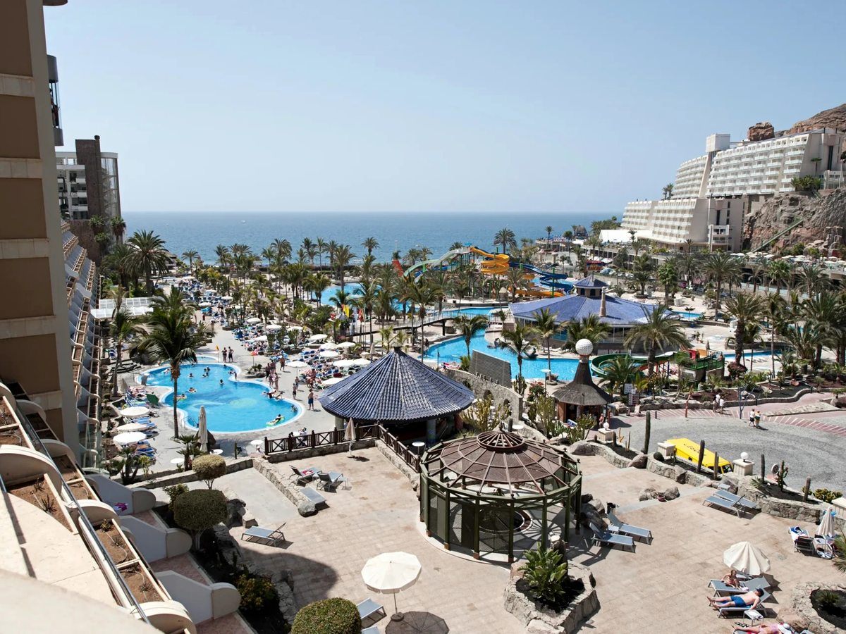 Elevated view over the Taurito valley: blue pools and sun terraces in the foreground, the waterpark's yellow and blue slides beyond, palms throughout and the Atlantic past a cliff-backed shore.