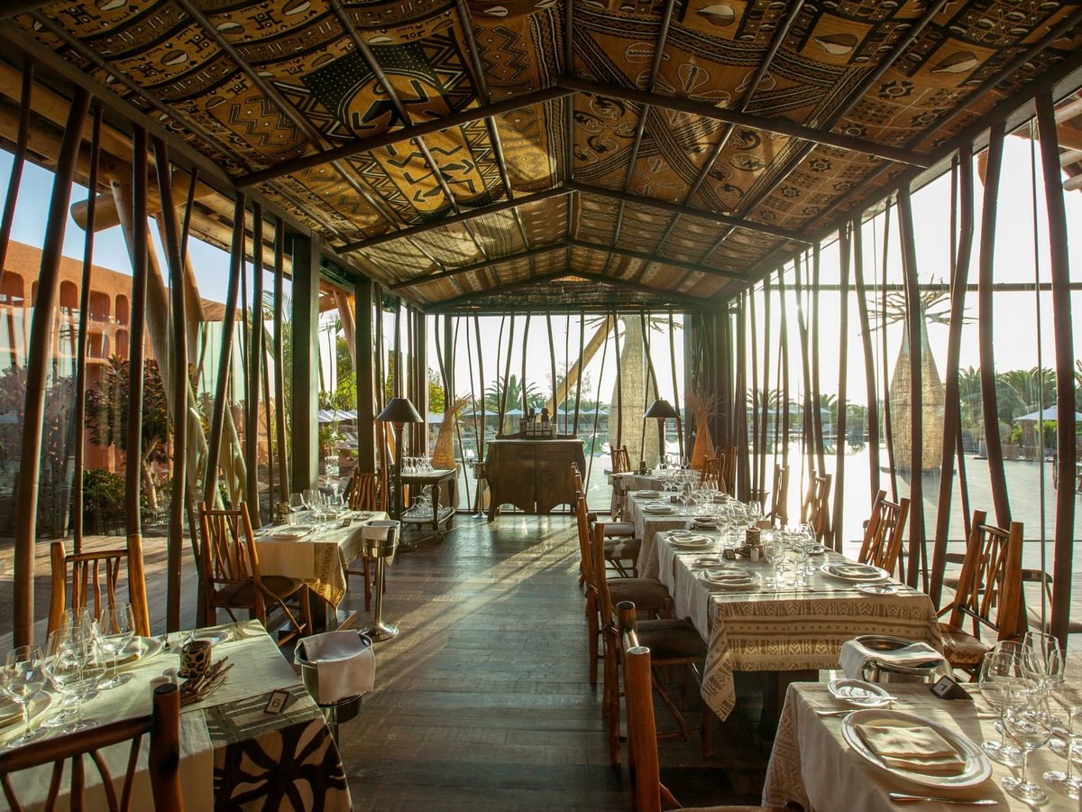 A glass-walled dining pavilion with a patterned fabric ceiling and slender timber poles, tables laid with white cloths and glassware, and the pool and gardens beyond the glass.