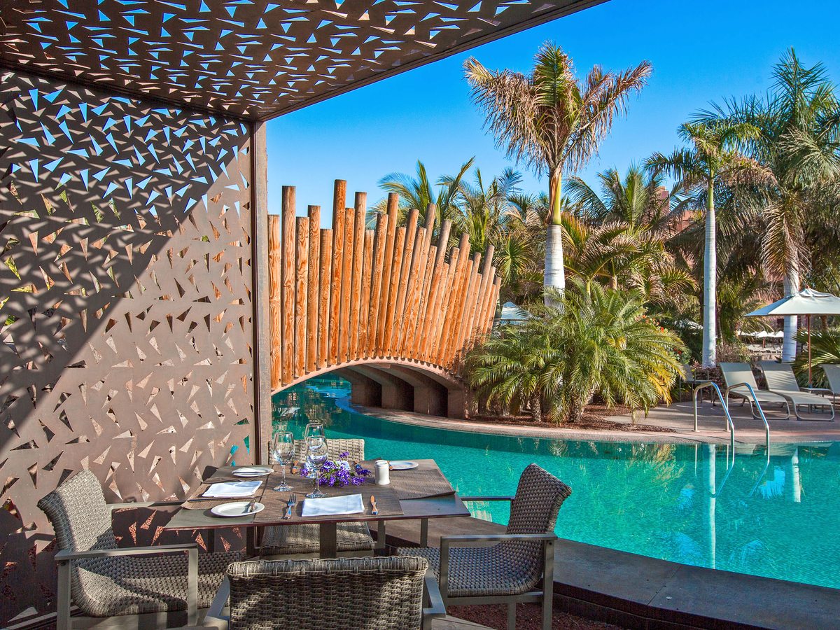 A laid table with wicker chairs on a shaded terrace at the edge of a turquoise pool, with a timber-poled bridge arching over the water and palms behind.