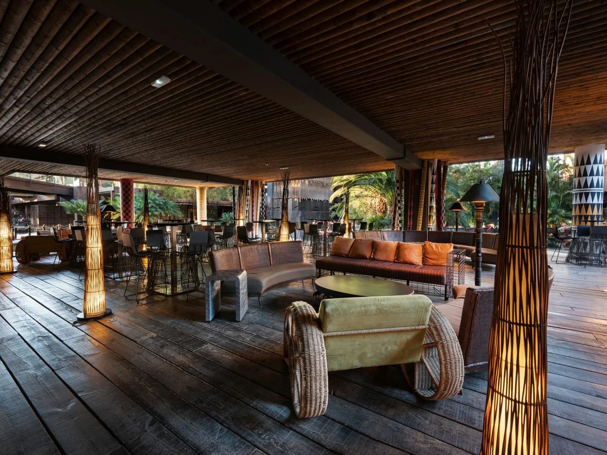 A covered open-air bar on timber decking with orange and olive sofas, low tables, bar stools at high tables and tall woven floor lamps, open to the gardens.