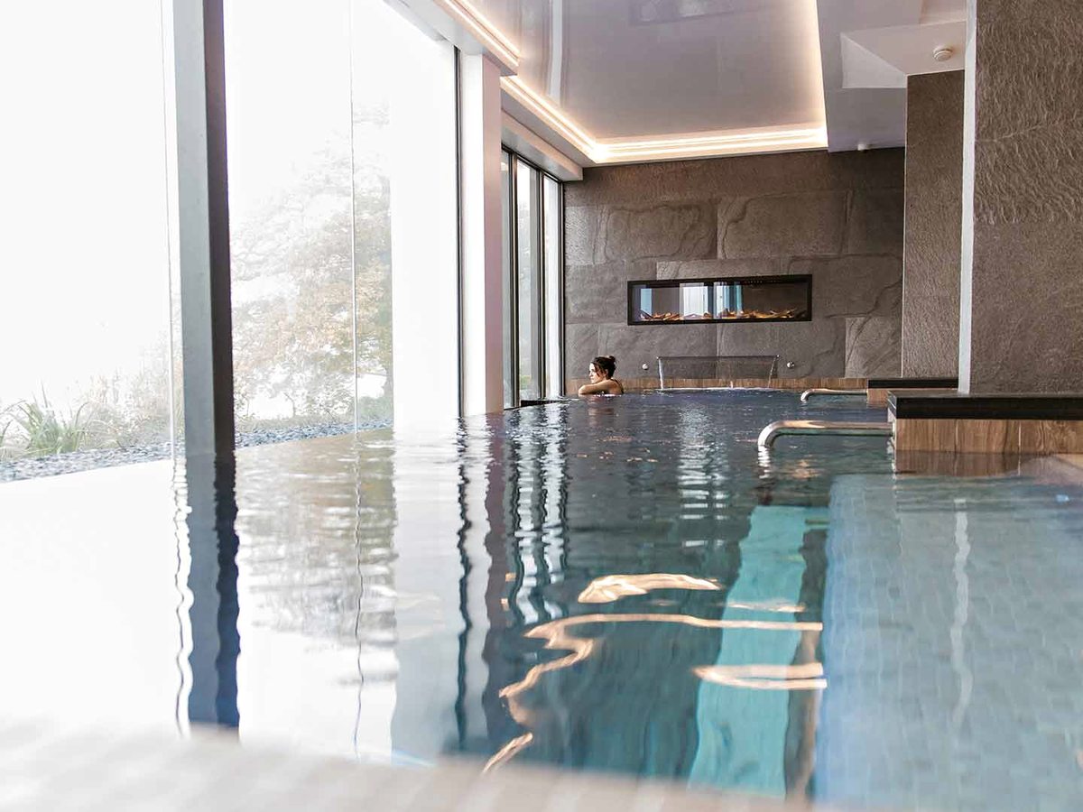 A dark-tiled indoor pool running alongside a full-height glass wall, with a stone-clad wall and a long fire feature at the far end.