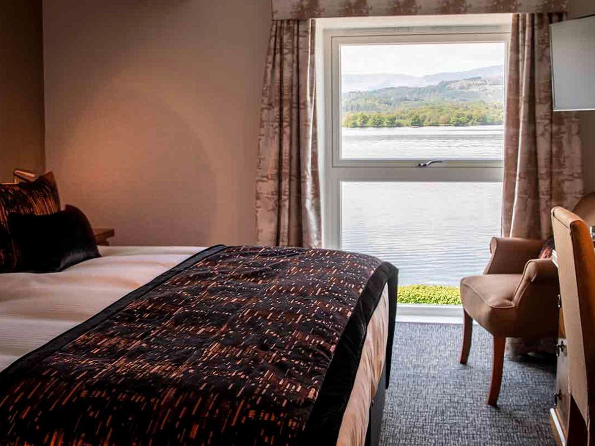 A bed with a copper-patterned throw beside a window framing the lake and the wooded far shore, with a leather armchair by the glass.