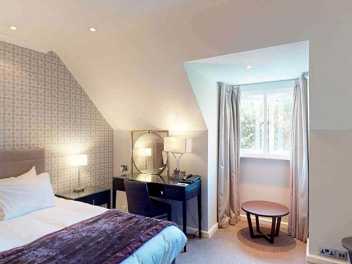 A bedroom with a sloping papered ceiling, a bed with a purple throw, a dark dressing table with a round mirror, and a small round table in the window recess.