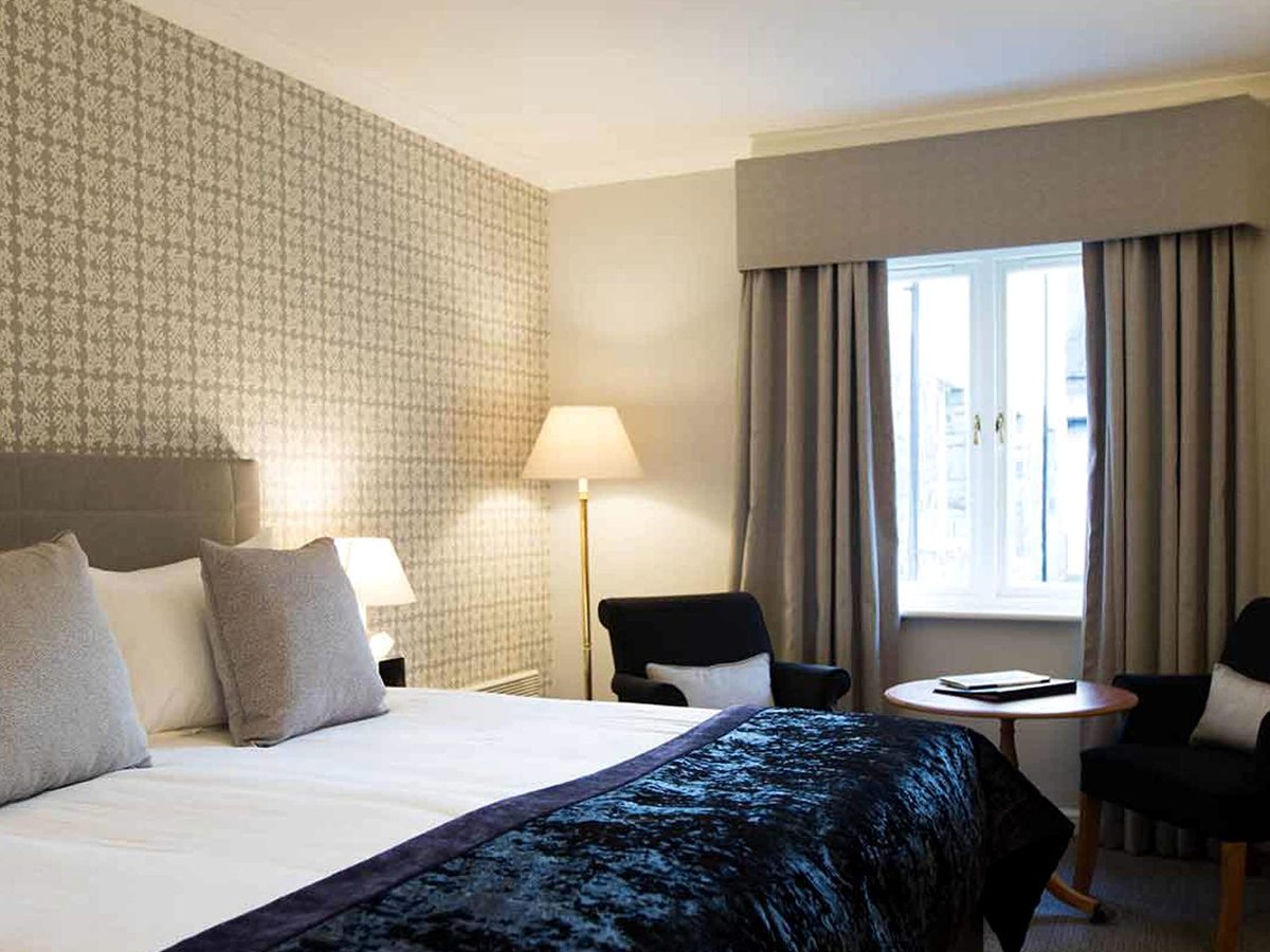 A bed with a dark blue velvet throw against a patterned papered wall, with a brass floor lamp and two dark chairs at a small round table by the window.