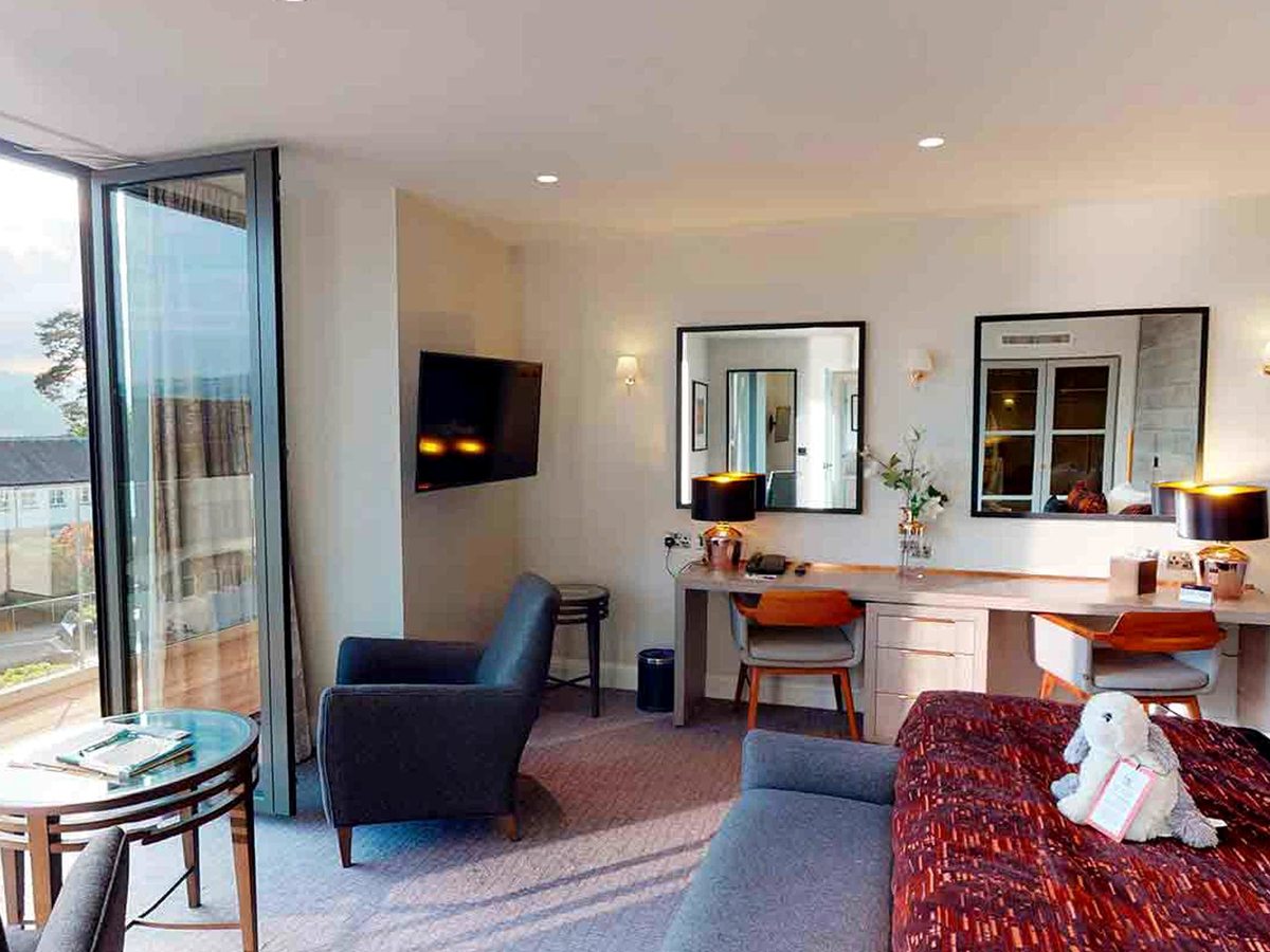 A room with twin dressing tables and mirrors, a wall-mounted television, a grey armchair and full-height glass doors onto a balcony.