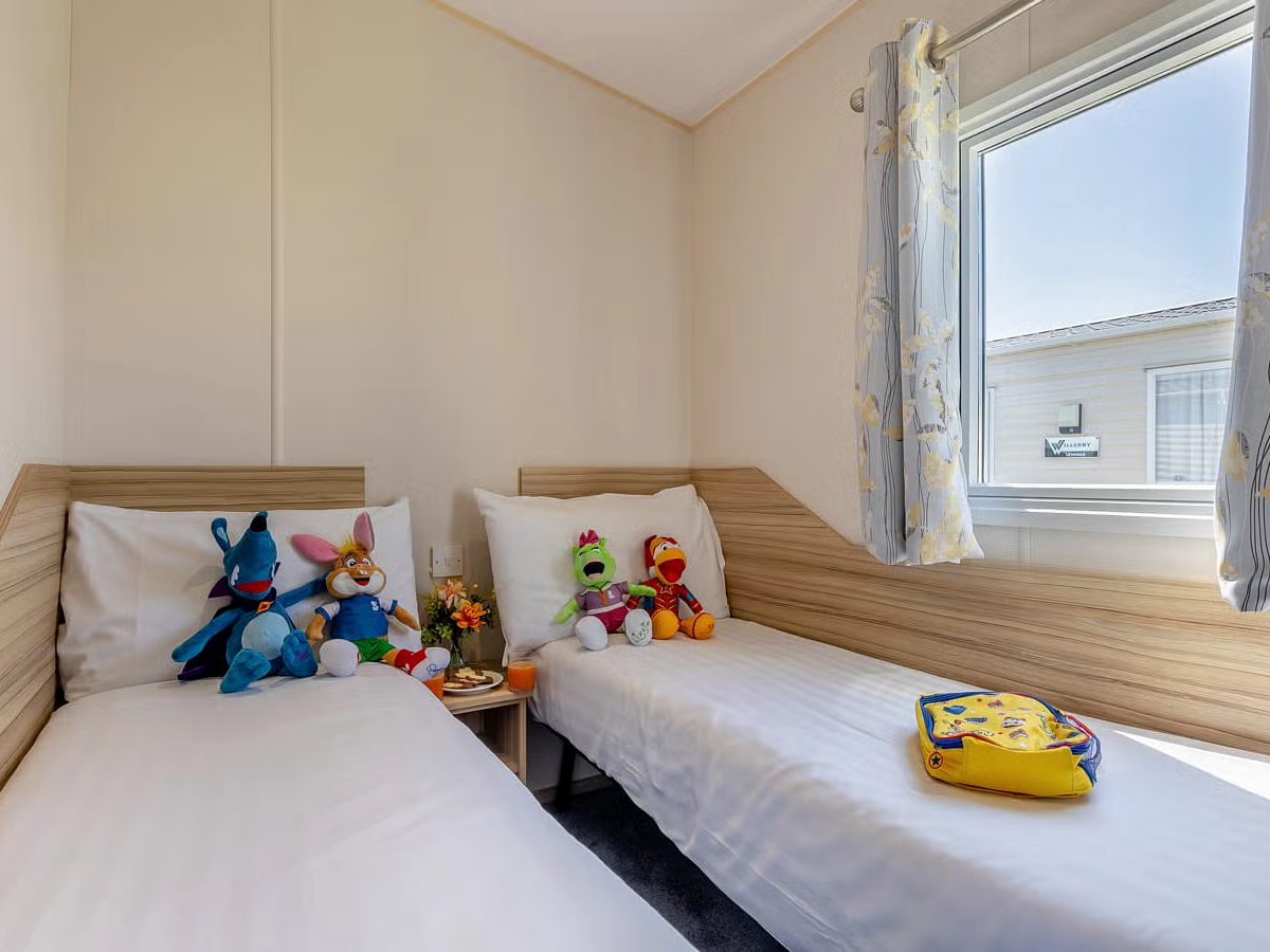 A caravan twin bedroom with two single beds side by side, white bedding, soft toys on the pillows and a curtained window between them.