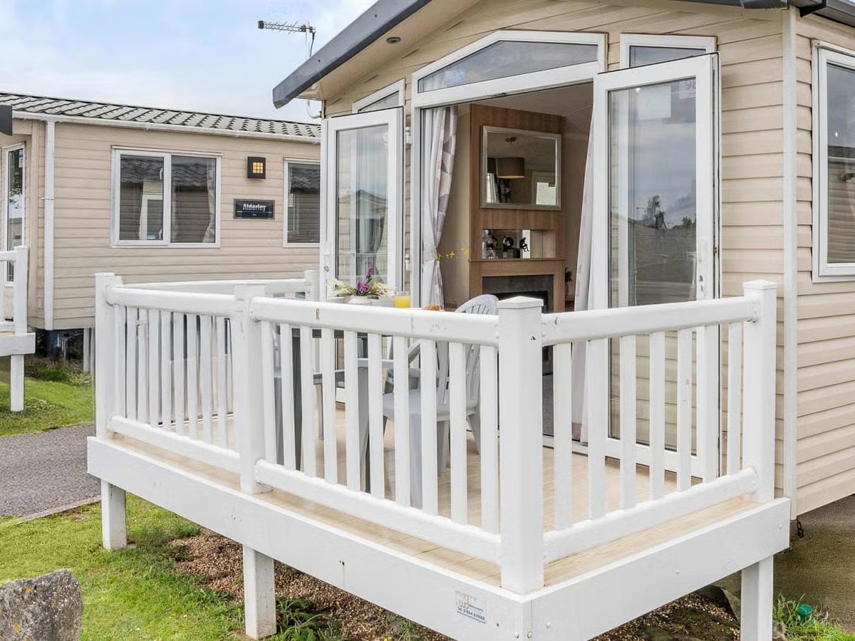 A cream static caravan with open French doors onto a raised timber deck enclosed by white railings, with a table and chairs on the deck.