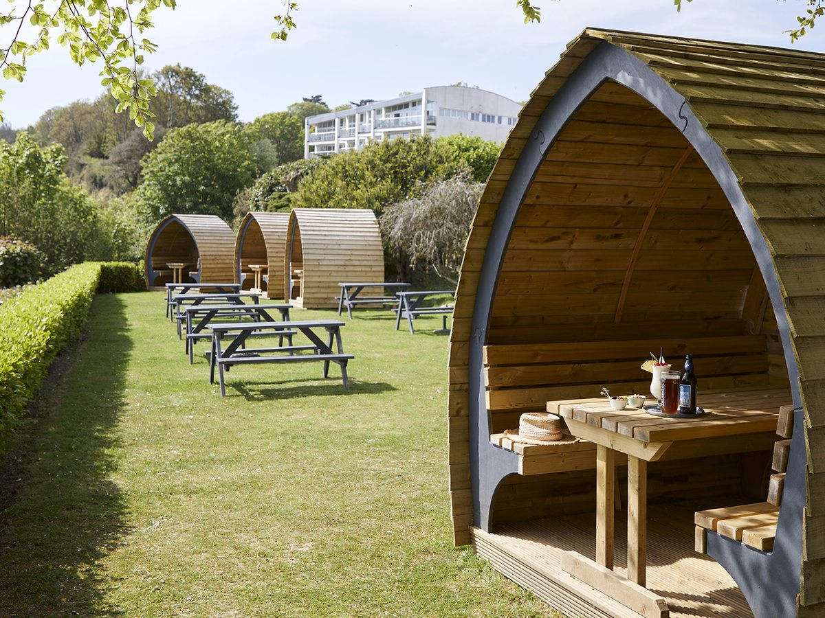 A row of curved wooden pods on a lawn, each with a picnic table inside, with more picnic tables out on the grass.