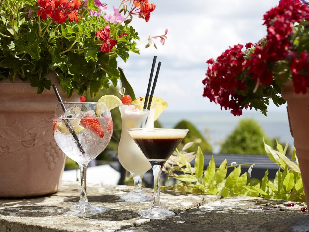 Three cocktails on a stone wall, with red geraniums in pots and the sea behind.
