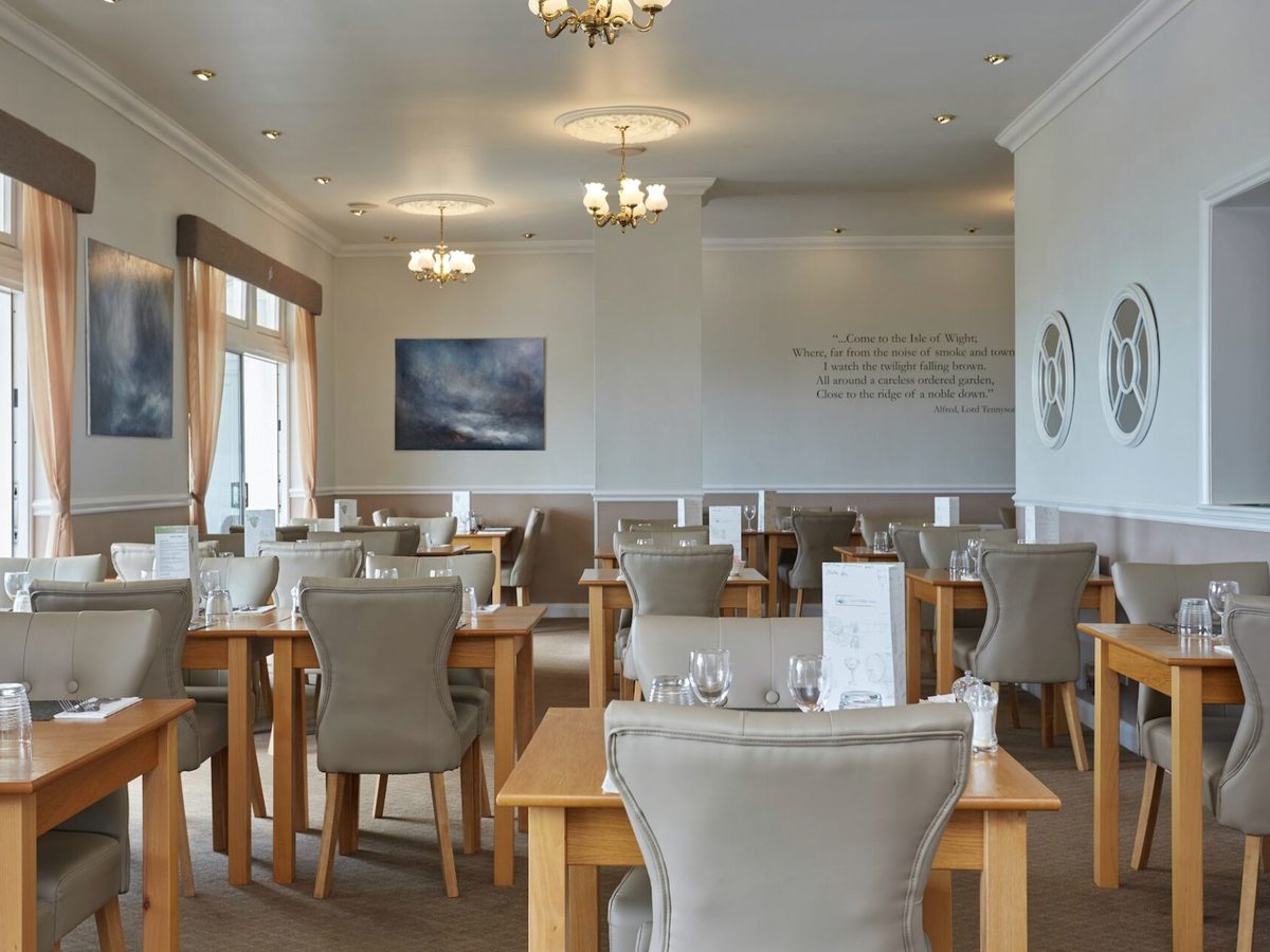 A light dining room with tables laid for dinner, upholstered chairs, tall windows and framed pictures on the walls.