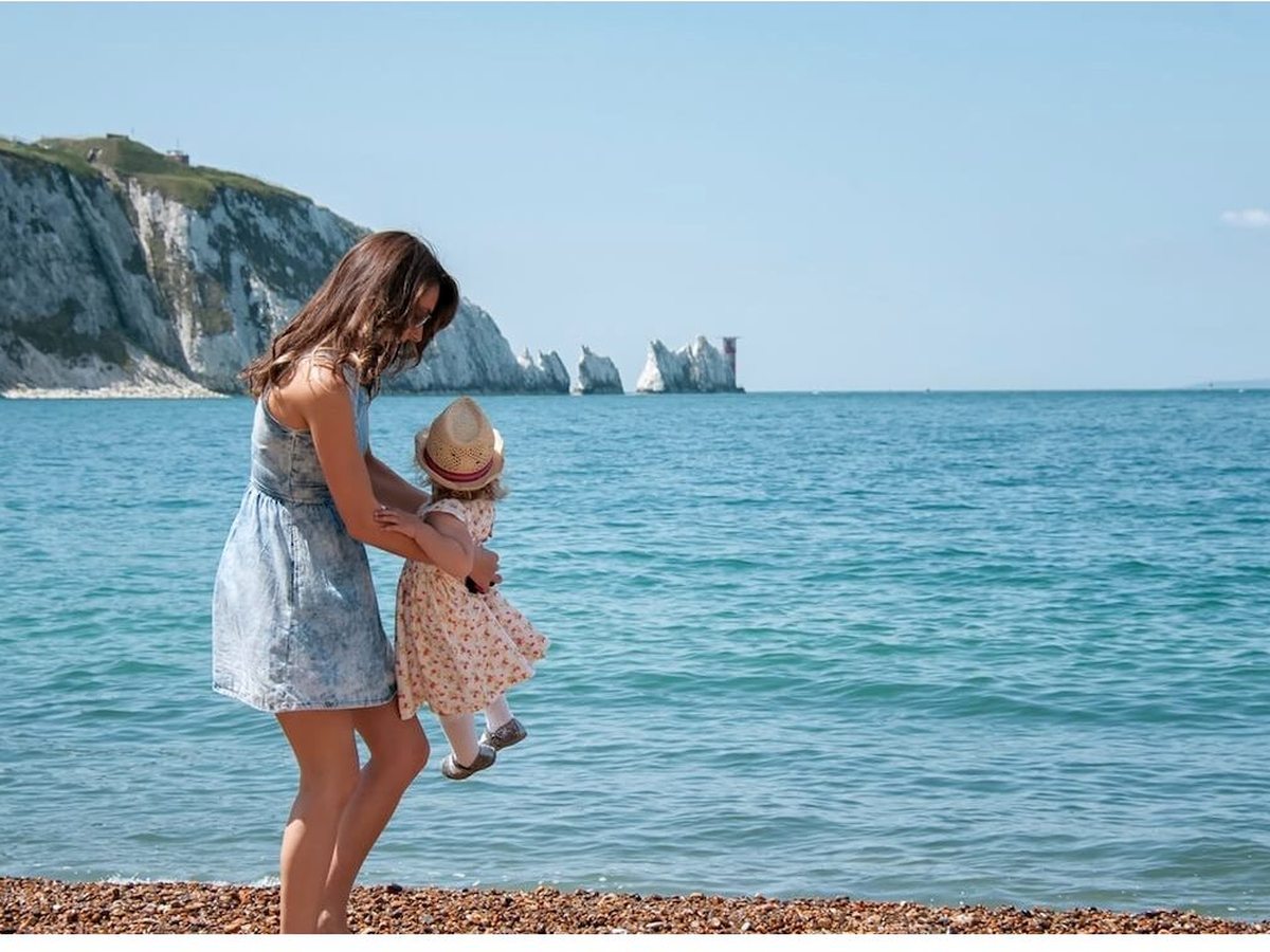 A woman lifting a toddler at the water's edge on a pebble beach, with white cliffs and the Needles stacks and lighthouse behind.