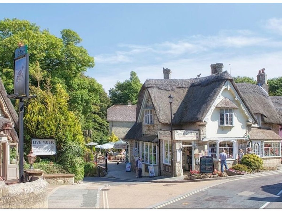 A thatched corner building with tearoom signs on a village street, tables under parasols and people walking past.