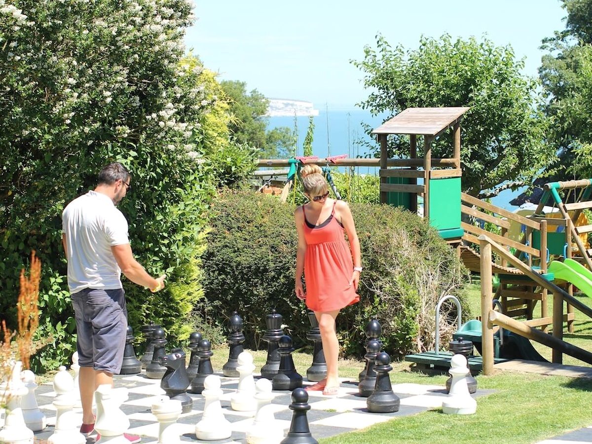 Two adults playing on a giant chess board on the lawn, with a wooden climbing frame and the sea behind them.
