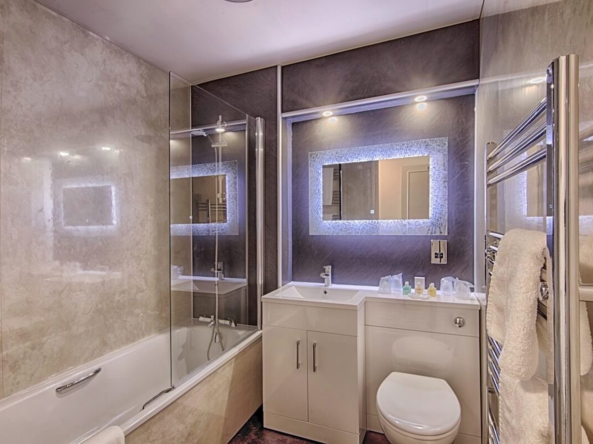 A bathroom with a bath and shower screen, a basin under a lit mirror and a heated towel rail.