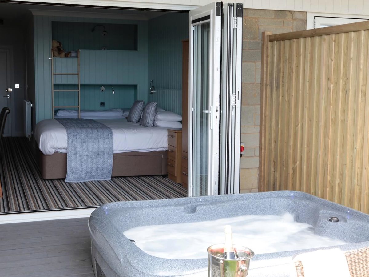 A bedroom with a double bed and built-in bunks, its folding doors open onto decking with a hot tub behind a screen.