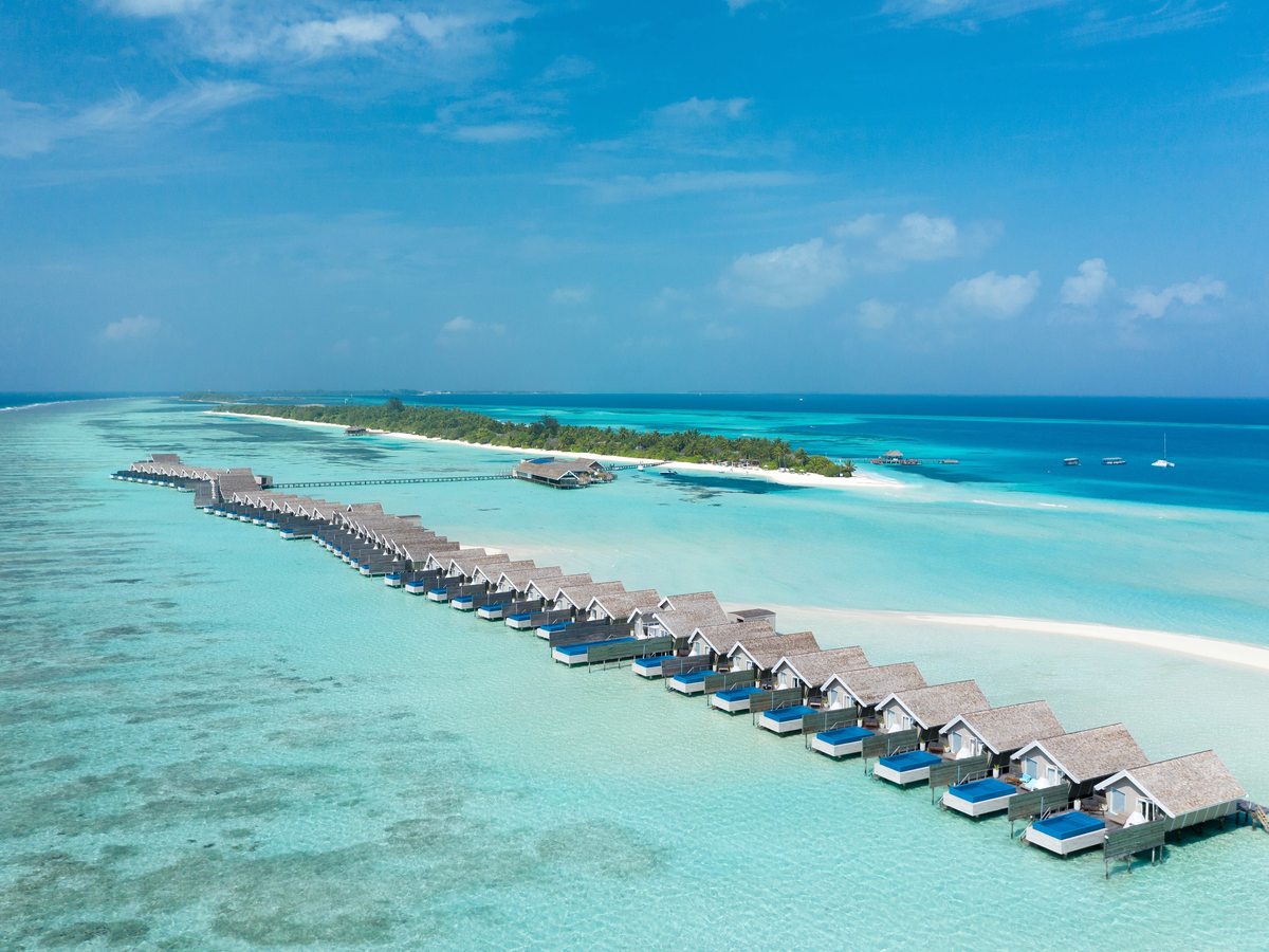 Aerial view of a line of thatched overwater villas with blue plunge pools stretching along the edge of a shallow turquoise lagoon, with the palm-covered island behind.