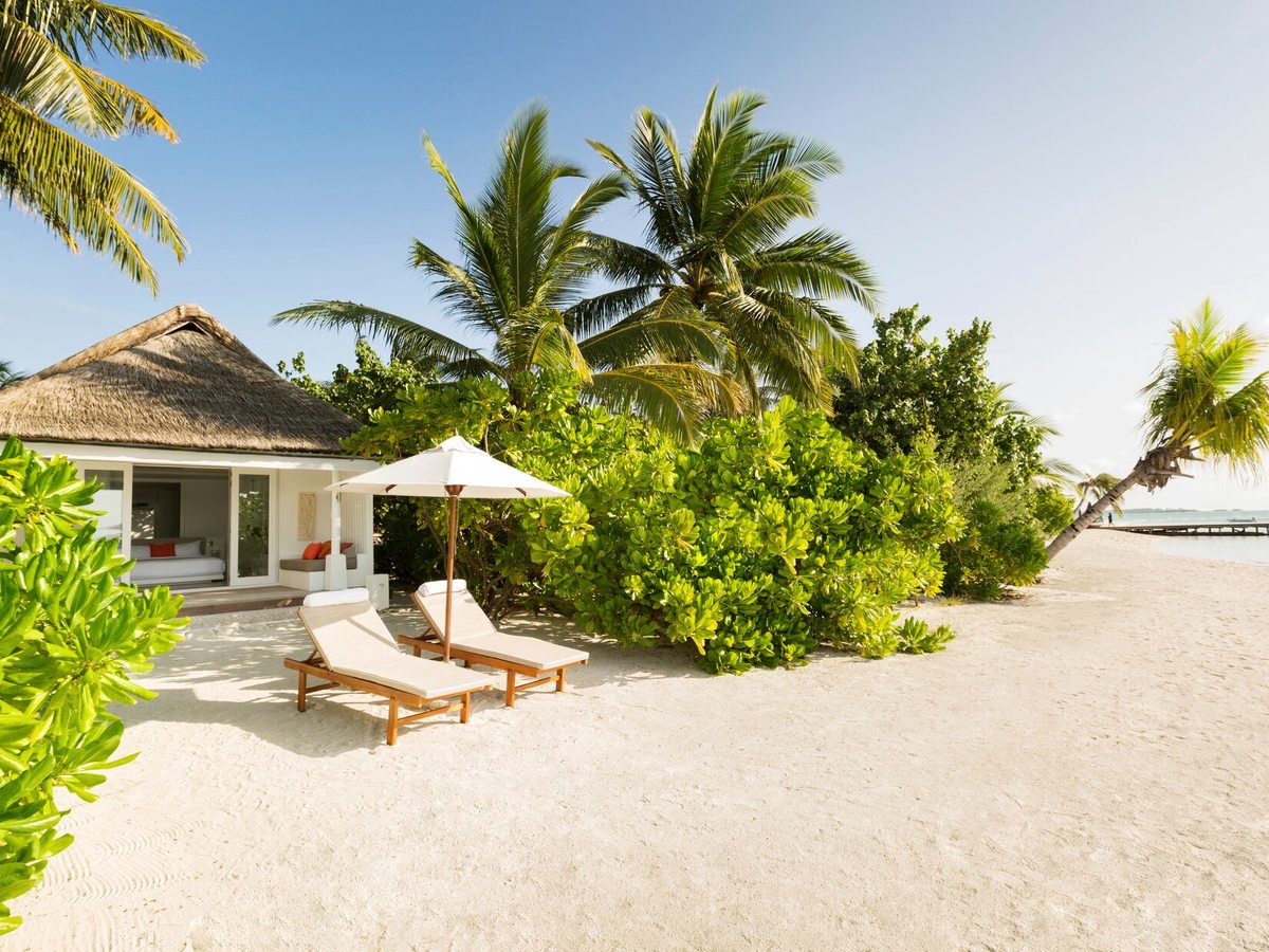 A thatched white villa among palms and low green scrub, with two wooden sun loungers and a parasol on the sand outside.