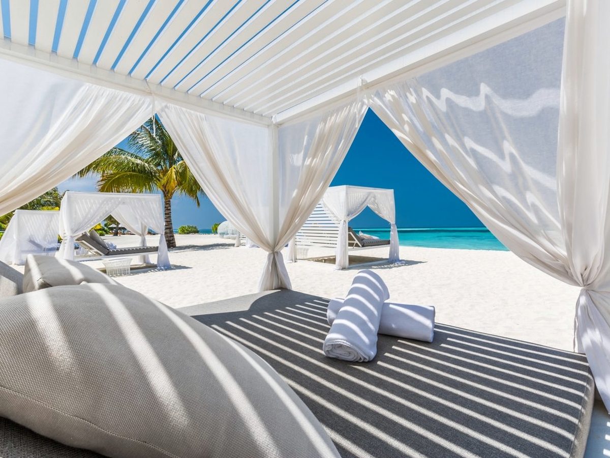 White curtained cabanas with cushioned daybeds and rolled towels set on white sand facing a bright blue lagoon.
