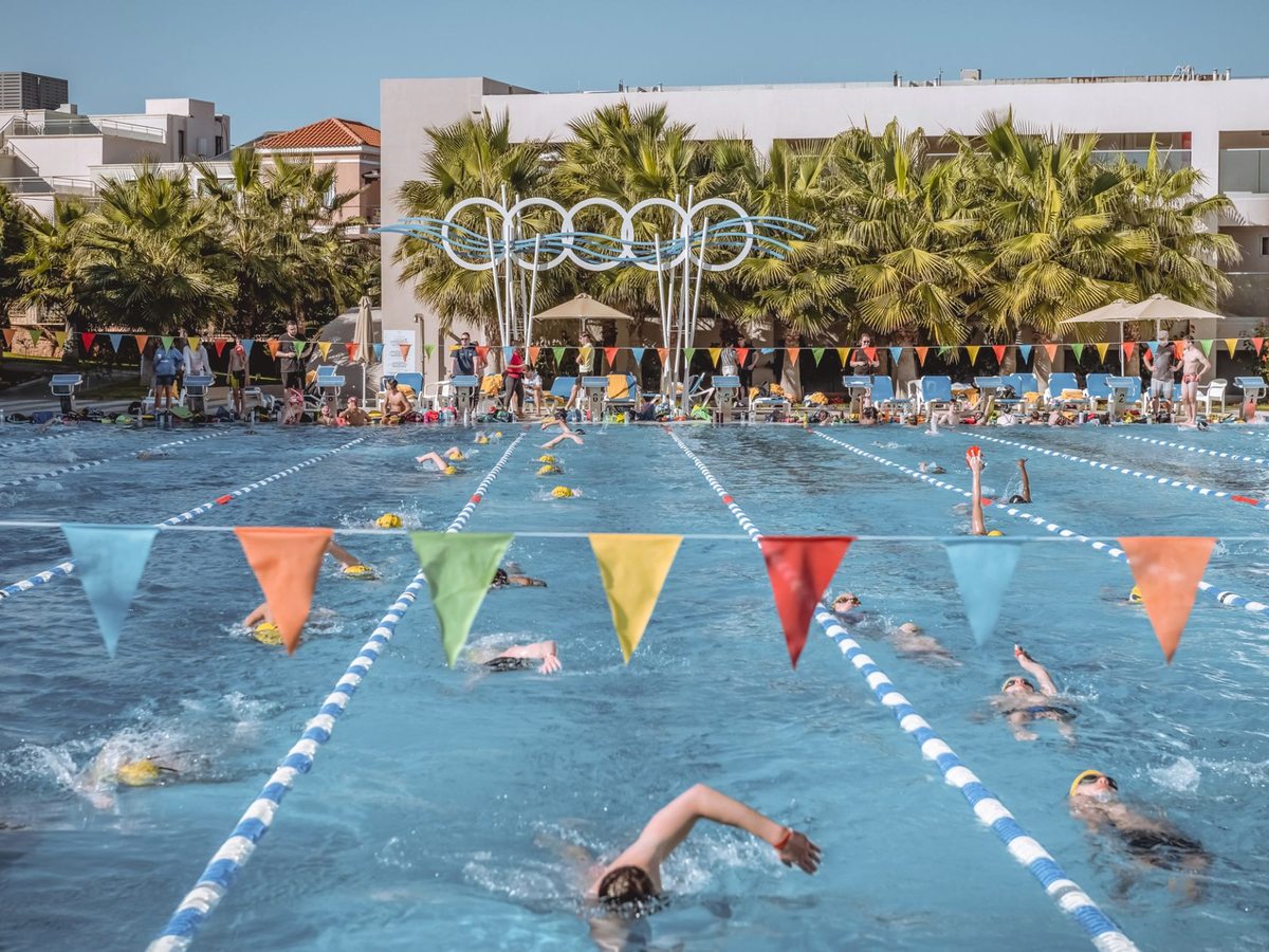 Swimmers doing lengths in a wide lane pool hung with triangular bunting, with palms and resort buildings behind.