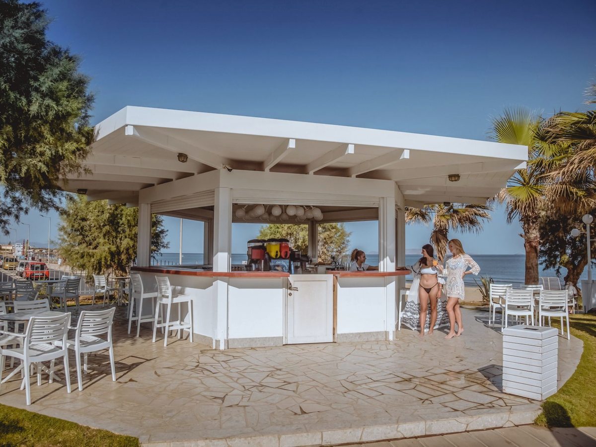 A white open-sided bar pavilion on a stone terrace by the lawn, with high stools, white tables and chairs and the sea behind.