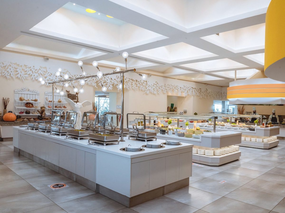 A large buffet hall with white serving islands of hot dishes and salads under bright ceiling lights.