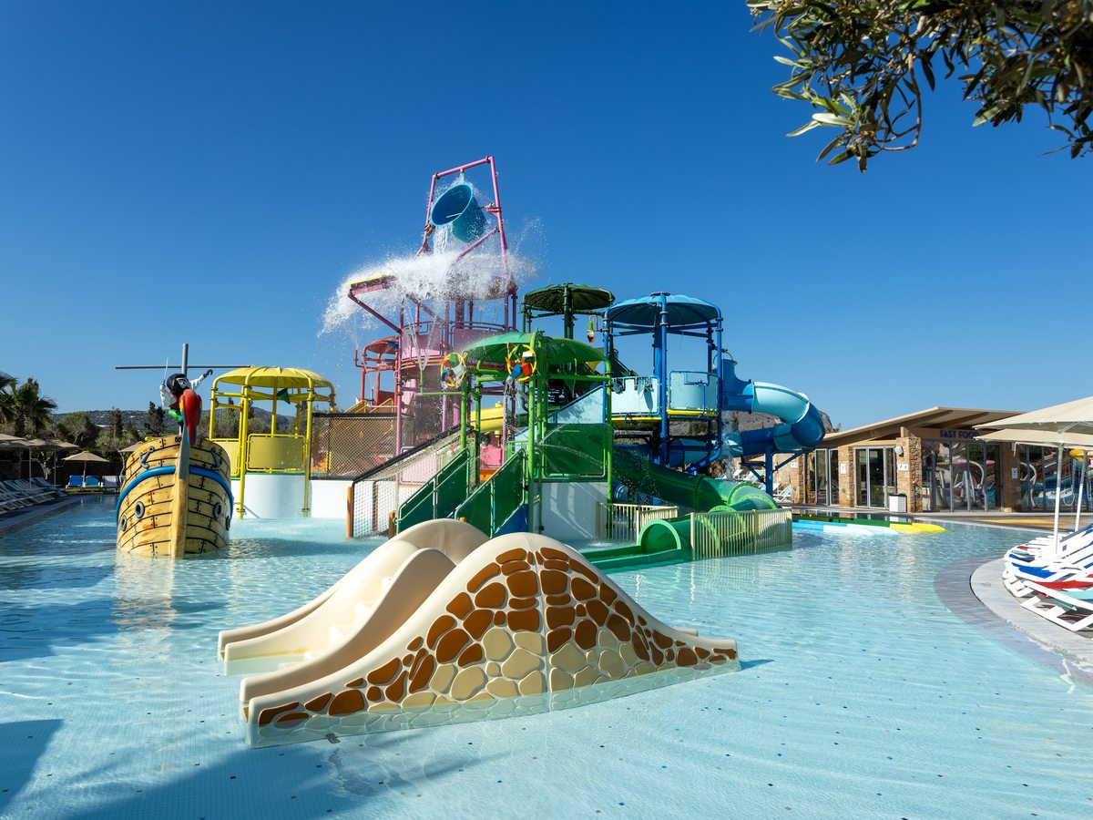 A multi-level children's water play structure with a wide slide and a tipping bucket standing in a shallow pool.