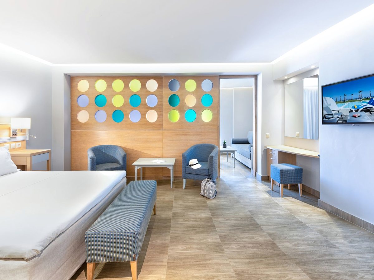 A suite with a bed and bench in the foreground, a headboard panel of coloured circles, and armchairs and a sofa in a sitting area beyond.