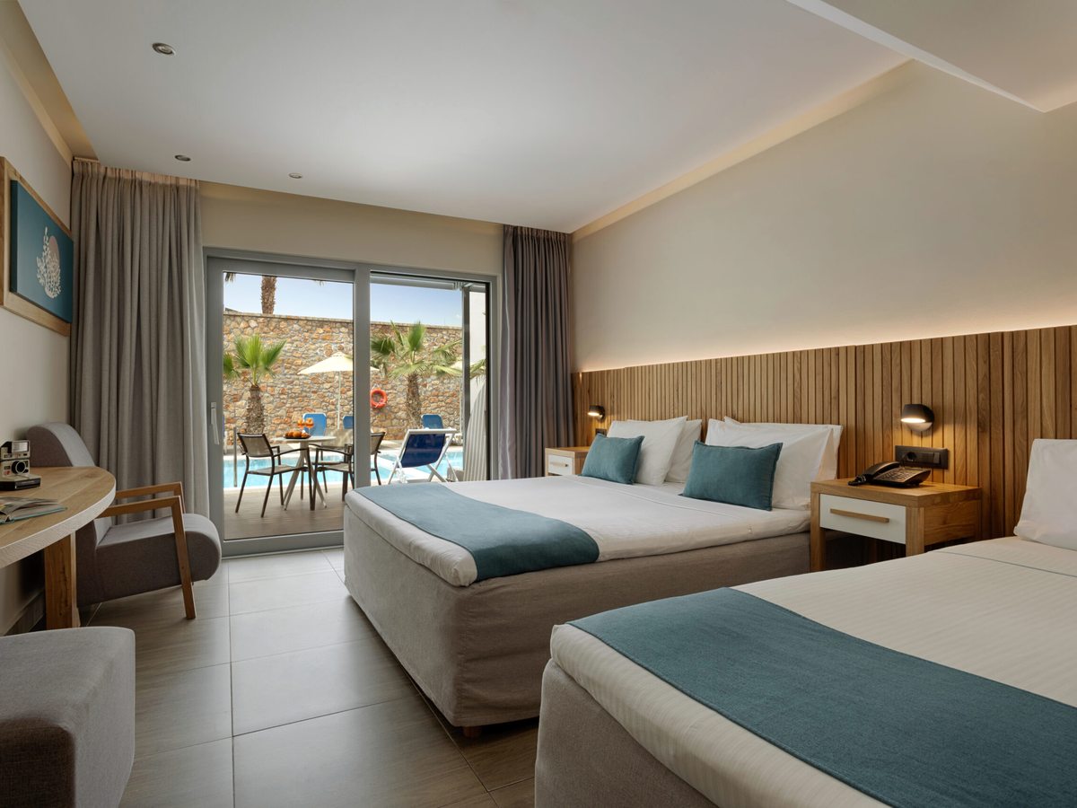 Twin beds with teal runners beside a desk, with glass doors open onto a terrace looking over palms and pool loungers.