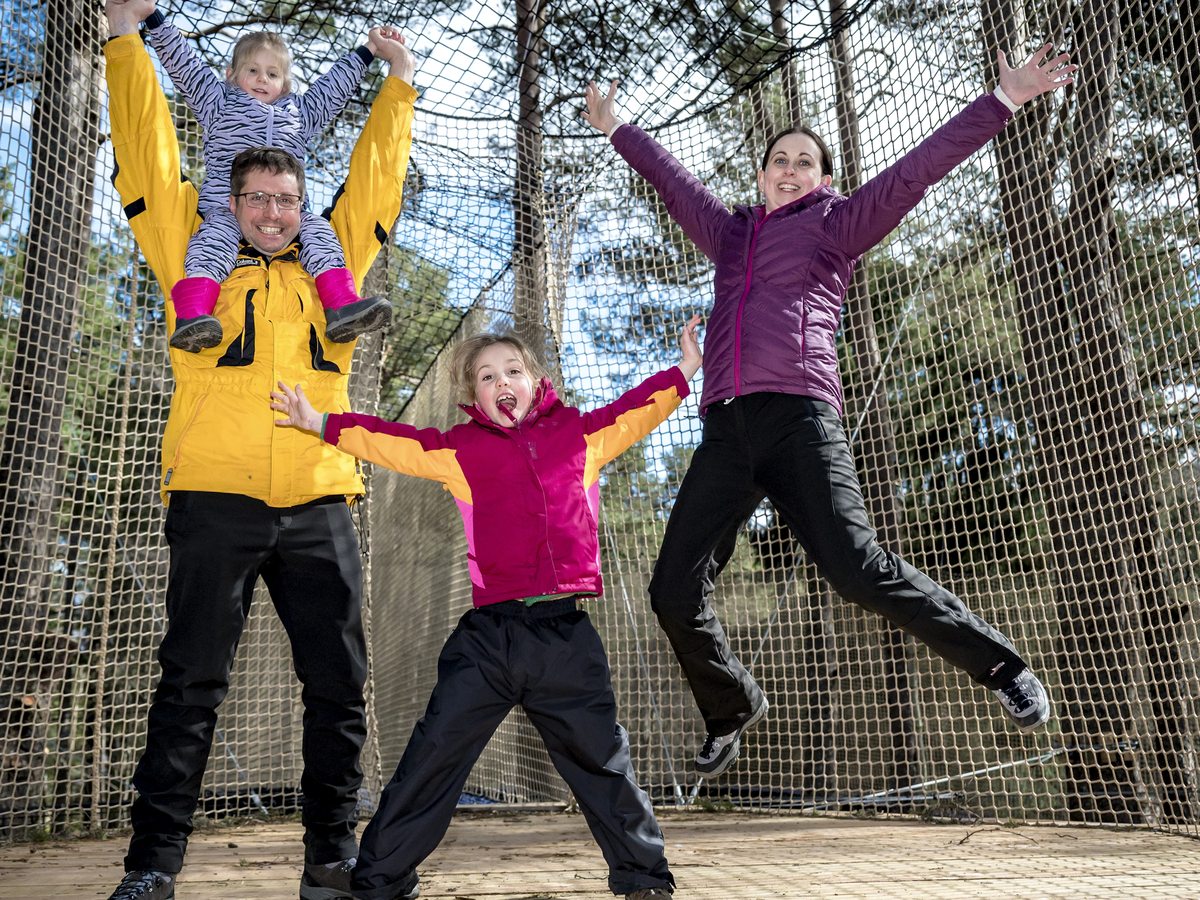 A family jumping in the air in front of a tall net enclosure strung between pine trees.