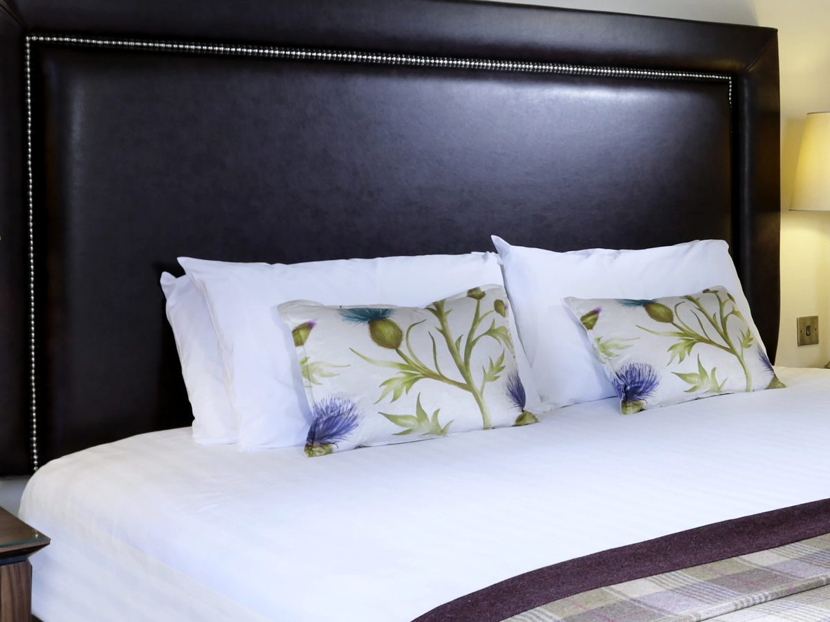 A double bed with crisp white bedding, thistle-print pillows and a tall dark leather headboard, with a tartan throw across the foot.