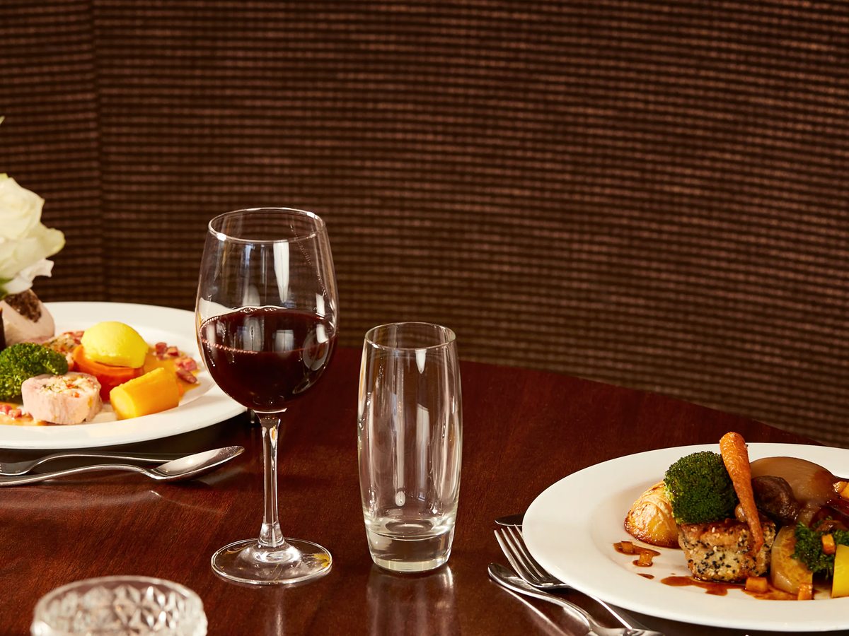 A restaurant table set with two plated main courses, a glass of red wine and a tumbler, in a booth panelled in dark wood.