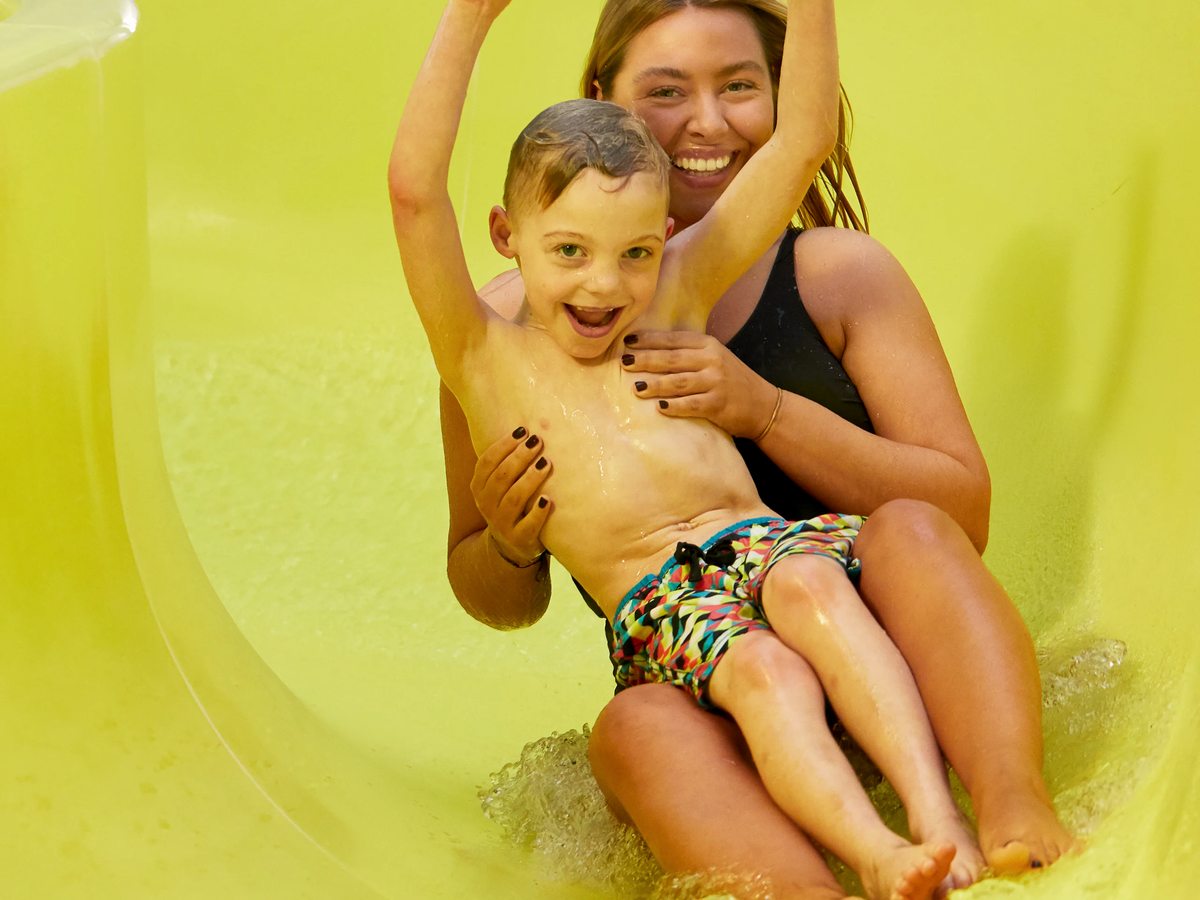 A woman and a young boy sliding down a bright yellow enclosed flume into shallow water.