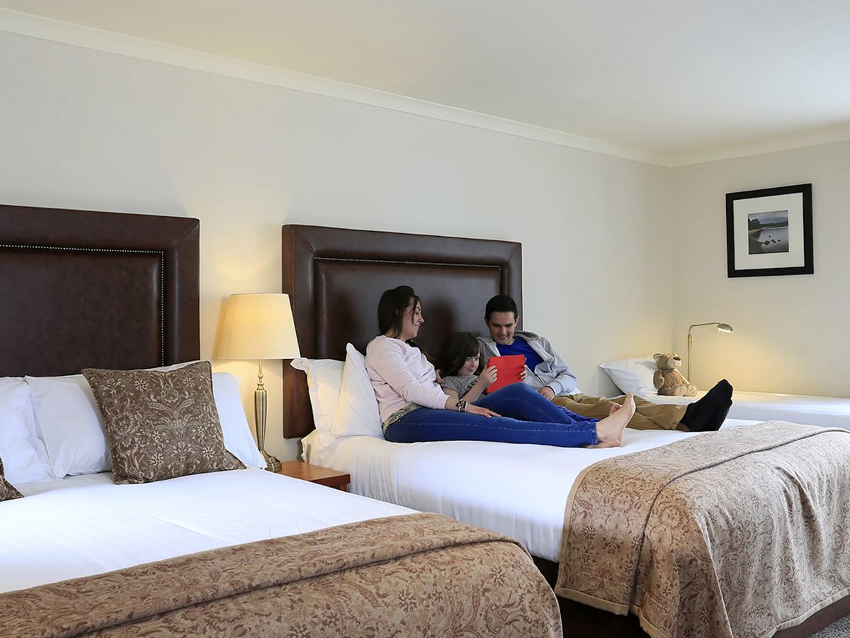 A family room with two large beds, dark padded headboards and bedside lamps, with a woman and a child sitting on one bed.