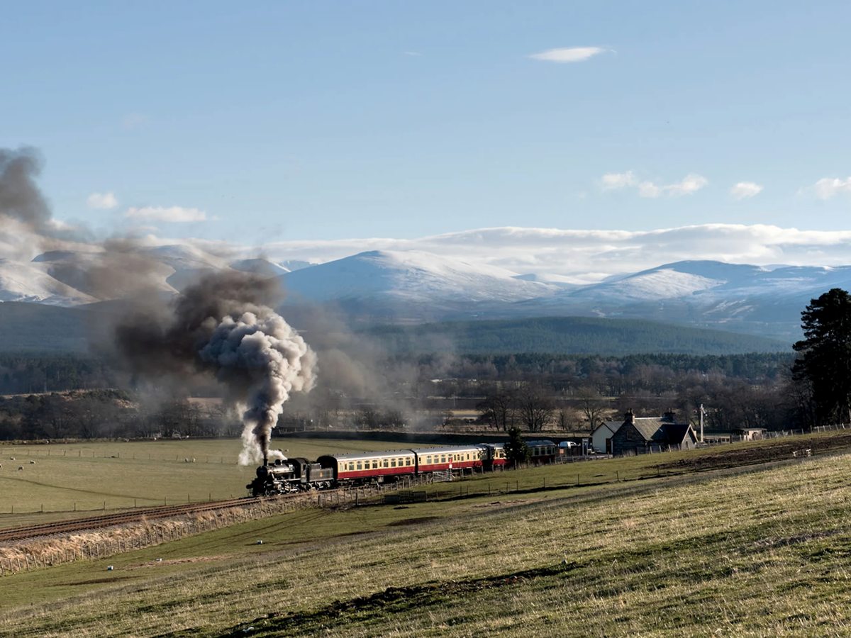 A steam locomotive hauling maroon and cream carriages across open fields, with a plume of dark smoke and snow-streaked mountains behind.